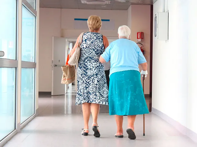 Zwei ältere Frauen gehen einen Krankenhausflur entlang, eine mit Gehstock.