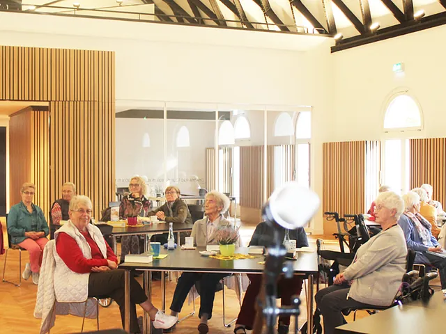 Gruppe von Senioren sitzt an Tischen in einem Raum.