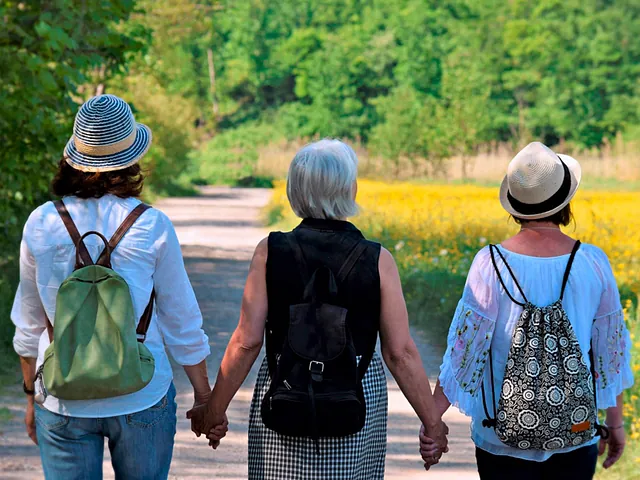 Drei Frauen mit Rucksäcken gehen Hand in Hand auf einem Weg im Grünen.